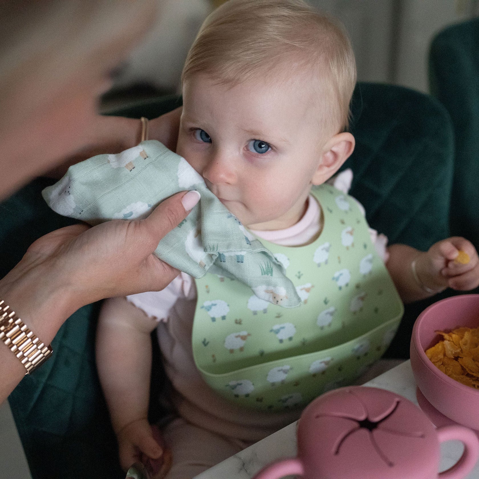 Baby wearing a green bib with sheep pattern, sitting at a table with a pink teapot and bowl.